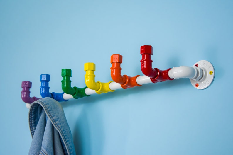 May include: A rainbow coloured pipe coat rack with a white pipe base. The rack is mounted to a light blue wall. A blue denim jacket is hanging on the rack.