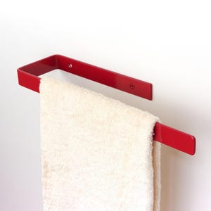 Può includere: Un portasciugamani in metallo rosso montato su una parete bianca, che tiene un asciugamano bianco piegato. Il portasciugamani ha un design rettangolare con una barra orizzontale e un supporto verticale. Le viti fissano il portasciugamani alla parete.