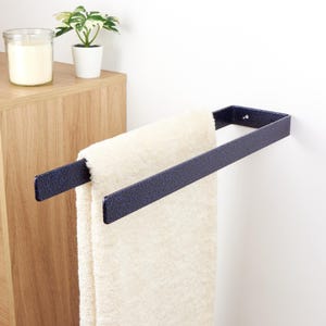 Può includere: Un portasciugamani in metallo blu navy montato su una parete bianca, che tiene un asciugamano color crema. Un mobile in legno e una piccola pianta in vaso sono visibili sullo sfondo. Il portasciugamani è largo circa 30 cm.