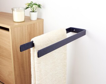 May include: A navy blue metal towel rack mounted on a white wall, holding a cream-colored towel. A wooden cabinet and a small potted plant are visible in the background. The towel rack is approximately 12 inches wide.