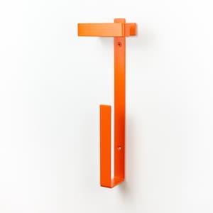 Op de afbeelding: Fel oranje metalen wandtoiletpapierhouder. De houder heeft een rechthoekig bovendeel, een lange verticale balk en een onderste haak voor het vasthouden van de toiletpapierrol. Gemonteerd op een witte muur.