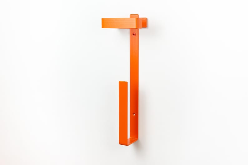 Op de afbeelding: Fel oranje metalen wandtoiletpapierhouder. De houder heeft een rechthoekig bovendeel, een lange verticale balk en een onderste haak voor het vasthouden van de toiletpapierrol. Gemonteerd op een witte muur.