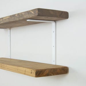 May include: Two wooden floating shelves with a natural finish are mounted on a white wall. The shelves are supported by white metal brackets. The top shelf has a darker stain than the bottom shelf. The shelves are designed for home decor and storage.