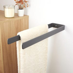 Può includere: Un portasciugamani in metallo grigio scuro montato su una parete bianca, che tiene un asciugamano color crema. Il portasciugamani ha un design semplice e moderno. Un mobile in legno e oggetti decorativi sono sullo sfondo.