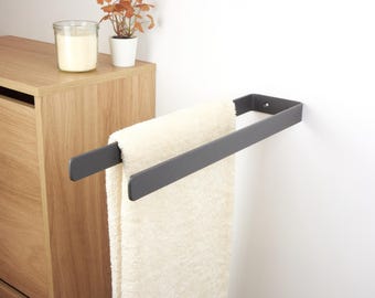 May include: A dark gray metal towel rack mounted on a white wall, holding a cream-colored towel. The rack has a simple, modern design. A wooden cabinet and decorative items are in the background.