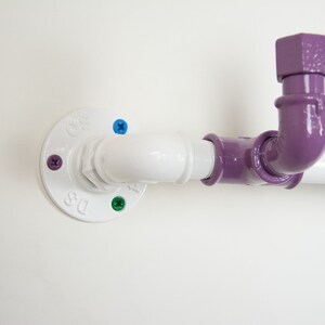 May include: A white pipe fitting with a purple pipe elbow and a blue pipe fitting. The pipe fitting is attached to a white wall with screws.