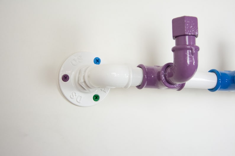 May include: A white pipe fitting with a purple pipe elbow and a blue pipe fitting. The pipe fitting is attached to a white wall with screws.