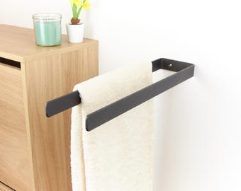 May include: A matte black towel rack mounted on a white wall, holding a cream-colored towel. The rack is rectangular and made of metal. A wooden cabinet and decorative items are visible in the background.