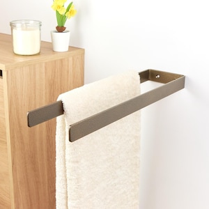 Könnte beinhalten: Ein bronzefarbener Handtuchhalter an einer weißen Wand, der ein gefaltetes cremefarbenes Handtuch hält. Ein Holzschrank und eine kleine Topfpflanze mit gelben Blumen sind im Hintergrund zu sehen.