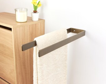 May include: A bronze-colored towel rack mounted on a white wall, holding a folded cream-colored towel. A wooden cabinet and a small potted plant with yellow flowers are visible in the background.