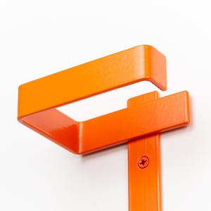 Op de afbeelding: Fel oranje metalen beugel met een rechthoekig ontwerp. De beugel is gemonteerd op een wit oppervlak en vastgezet met een schroef. Het ontwerp heeft een bovendeel en een verticale steun.
