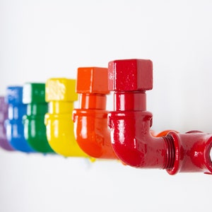 May include: A row of colourful pipe fittings painted in rainbow colours, starting with purple, blue, green, yellow, orange, and ending with red. The pipe fittings are mounted on a white wall.