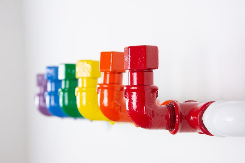 May include: A row of colourful pipe fittings painted in rainbow colours, starting with purple, blue, green, yellow, orange, and ending with red. The pipe fittings are mounted on a white wall.