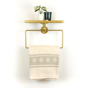 Könnte beinhalten: Goldfarbenes Wandregal mit Handtuchhalter. Ein cremefarbenes Handtuch mit Blättermuster hängt am Halter. Zwei kleine Topfpflanzen stehen auf dem Regal. Die Halterung ist an einer weißen Wand befestigt.