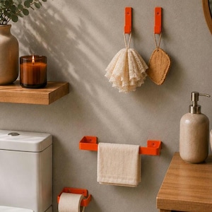 Può includere: Un set di accessori da bagno color arancione brillante composto da due ganci a parete con una spugna a rete beige e una spugna abrasiva, un portasciugamani con un asciugamano beige piegato e un portarotolo con carta igienica bianca.