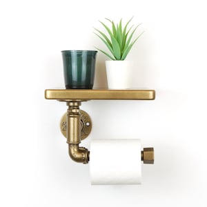 May include: A wall-mounted industrial-style toilet paper holder with a shelf. The holder is made of brass-colored metal pipes and fittings. A roll of white toilet paper is in place. A small green glass and a potted plant sit on the shelf.