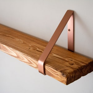 May include: A wooden shelf with a copper metal bracket. The shelf is made of natural wood with a rustic finish. The bracket is a simple triangle shape.