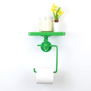 Op de afbeelding: Een heldergroene wandplank en toiletpapierhouder. De plank bevat een kaars in een glazen pot en een kleine pot met gele narcissen. De toiletpapierhouder bevindt zich onder de plank en bevat een rol wit papier.