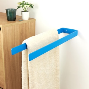 Könnte beinhalten: Ein moderner, blauer Metall-Handtuchhalter an einer weißen Wand, der ein cremefarbenes Handtuch hält. Der Halter hat ein einfaches, rechteckiges Design. Ein Holzschrank und Topfpflanzen befinden sich im Hintergrund.