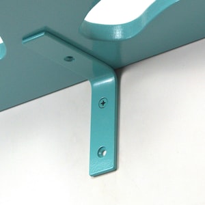 May include: A teal wooden shelf with a decorative cutout design. The shelf is supported by a matching teal metal bracket, attached to a white wall with screws. The bracket is L-shaped and has pre-drilled holes for mounting.