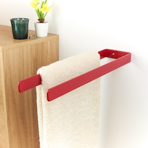 Può includere: Un portasciugamani in metallo rosso montato su una parete bianca, che tiene un asciugamano color crema piegato. La barra ha un design semplice e moderno. Un mobile in legno e oggetti decorativi sono sullo sfondo.