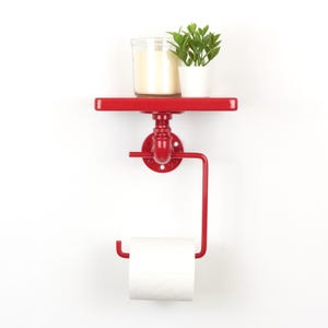 Può includere: Mensola da parete in metallo rosso con portarotolo integrato. La mensola contiene una candela in un barattolo di vetro e una piccola pianta in vaso. Il design è ispirato ai raccordi idraulici.