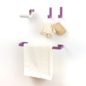 Può includere: Un set da bagno viola con un portasciugamani, due ganci e un portarotolo. Un asciugamano bianco è appeso al portasciugamani, e una spugna vegetale e un guanto da bagno sono appesi ai ganci.