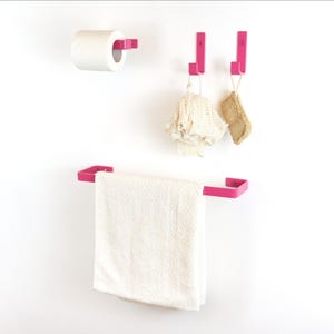 Può includere: Un set di accessori per il bagno rosa acceso contro un muro bianco. Il set comprende un portarotolo, due ganci e un portasciugamani. Un asciugamano bianco è appeso al portasciugamani. Un guanto da bagno bianco e una spugna marrone sono appesi ai ganci.