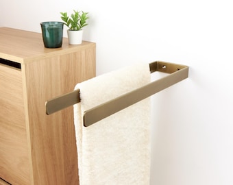 May include: A brass-colored towel rack mounted on a white wall, holding a folded cream-colored towel. The rack is rectangular and has a minimalist design. A wooden cabinet and small potted plants are visible in the background.
