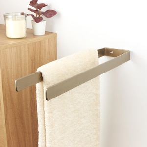 Könnte beinhalten: Ein moderner Handtuchhalter aus gebürstetem Metall, montiert an einer weißen Wand, mit einem cremefarbenen Handtuch. Ein Holzschrank und eine kleine Topfpflanze sind im Hintergrund zu sehen. Der Handtuchhalter ist etwa 30 cm breit.