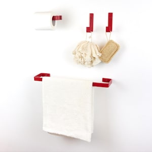 Può includere: Un set di accessori da bagno rossi contro un muro bianco. Include un portarotolo, due ganci e un portasciugamani. Un asciugamano bianco è appeso al portasciugamani. Due spugne da bagno sono appese ai ganci.