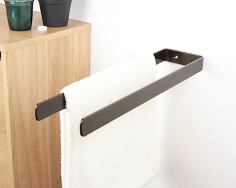 May include: A dark bronze towel bar mounted on a white wall, holding a folded white towel. The bar has a simple, modern design. A wooden cabinet and two small potted plants are visible in the background.