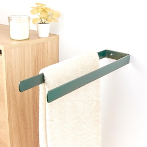 Può includere: Un moderno portasciugamani in metallo verde scuro montato su una parete bianca, che tiene un asciugamano color crema. Il portasciugamani ha un design semplice e minimalista. Un mobile in legno e oggetti decorativi sono sullo sfondo.