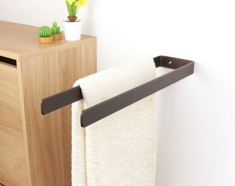 May include: A dark brown metal towel rack mounted on a white wall, holding a cream-colored towel. The rack has a simple, rectangular design. A wooden cabinet and small potted plants are visible in the background.