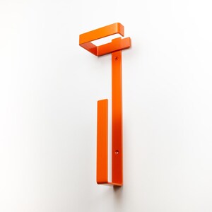 Op de afbeelding: Een oranje metalen wandkapstok. De kapstok heeft een rechthoekig bovendeel en een verticale balk met een onderste haak. Het ontwerp is minimalistisch en modern, geschikt voor entrees of slaapkamers.