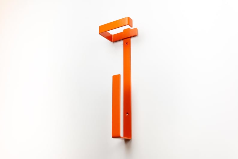 Op de afbeelding: Een oranje metalen wandkapstok. De kapstok heeft een rechthoekig bovendeel en een verticale balk met een onderste haak. Het ontwerp is minimalistisch en modern, geschikt voor entrees of slaapkamers.