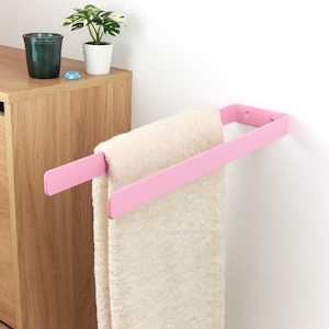 Può includere: Un portasciugamani in metallo rosa montato su una parete bianca, che tiene un asciugamano color crema. Il portasciugamani ha un design semplice e moderno. Un mobile in legno e piccole piante in vaso sono sullo sfondo.