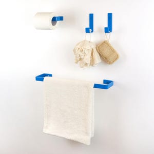 Può includere: Un set di accessori da bagno blu contro un muro bianco. Include un portarotolo di carta igienica, due ganci con una luffa e una spugna, e un portasciugamani con un asciugamano bianco. Design moderno e minimalista.
