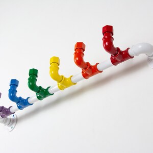 May include: A rainbow coloured pipe coat rack with a white pipe and a white wall mount. The rack has seven hooks, each a different colour of the rainbow.