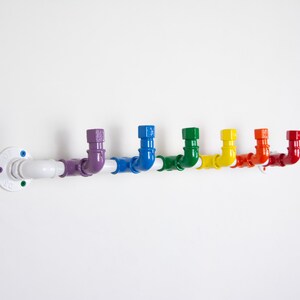 May include: A white metal pipe with rainbow coloured hooks. The pipe is mounted to a white wall. The hooks are in the shape of a 90-degree bend.