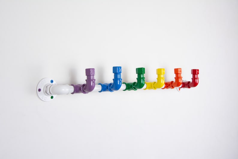 May include: A white metal pipe with rainbow coloured hooks. The pipe is mounted to a white wall. The hooks are in the shape of a 90-degree bend.