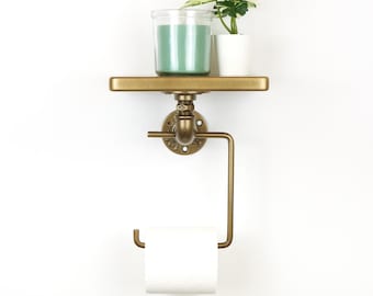 May include: A wall-mounted, industrial-style toilet paper holder with a shelf. The holder is made of brass-colored metal and features a shelf holding a green candle in a glass jar and a small potted plant. A roll of white toilet paper is in place.