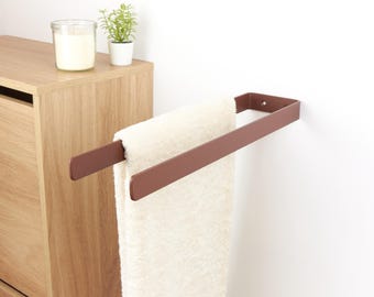 May include: A modern, brown metal towel rack mounted on a white wall, holding a folded white towel. The rack has a simple, rectangular design. A wooden cabinet, a candle in a glass jar, and a small potted plant are visible in the background.