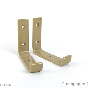 May include: Two champagne gold gloss metal shelf brackets with a hook on the end. The brackets are angled and have three holes for mounting.