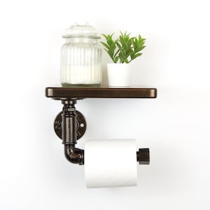 May include: A rustic, industrial-style bathroom shelf with a dark brown wooden shelf, a metal pipe toilet paper holder, and a small potted plant. A clear glass jar with a lid sits on the shelf. The toilet paper roll is white.