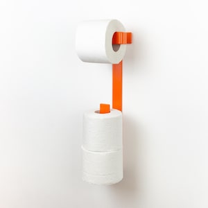 Op de afbeelding: Een oranje metalen toiletpapierhouder aan een witte muur. De houder heeft een verticale stang met een boven- en ondergedeelte om twee rollen wit toiletpapier te bevatten. Het ontwerp is modern en minimalistisch.