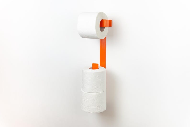 Op de afbeelding: Een oranje metalen toiletpapierhouder aan een witte muur. De houder heeft een verticale stang met een boven- en ondergedeelte om twee rollen wit toiletpapier te bevatten. Het ontwerp is modern en minimalistisch.