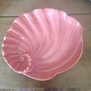 May include: A pink ceramic seashell-shaped bowl with a detailed spiral design. The bowl is perfect for serving snacks or holding decorative items.