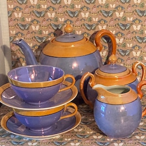 May include: A vintage tea set in shades of blue and orange. The set includes a teapot, creamer, sugar bowl, and two teacups with saucers. The tea set has gold trim and handles. The background features a butterfly pattern.
