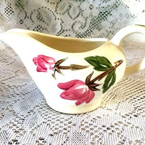 May include: Cream-colored gravy boat with a handle, featuring pink rose and green leaf floral designs. The gravy boat is made of ceramic and is likely vintage.